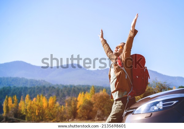 Man with arms raised up
cheerful and happy sitting on car. Tourist with backpack enjoy
mountain views.