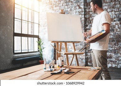 Man Artist Stands In Front Of A Blank Canvas On Easel With Palette In Loft Art Studio. Male Painter Hold Paintbrush In Hand And Mix Paint. Hobby Concept