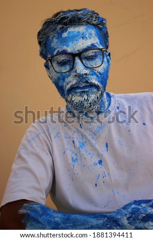 Man artistically covered in blue and white oil paint, front profile with glasses on brown blurry background. Making it a classy yet unique wallpaper.