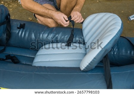 A man assembling a blue inflatable kayak outdoors, his hands attaching the seat with a black strap, concept of active recreation, sports equipment preparation, summer hobbies. 