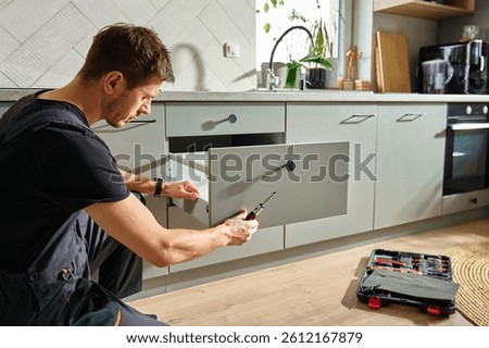 Man assembling kitchen drawer in modern cabinet using screwdriver. Handyman installing and adjusting drawer slide mechanism in contemporary kitchen. Furniture installation