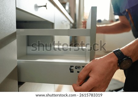 Man assembling kitchen drawer in modern cabinet using screwdriver. Handyman installing and adjusting drawer slide mechanism in contemporary kitchen. Furniture installation