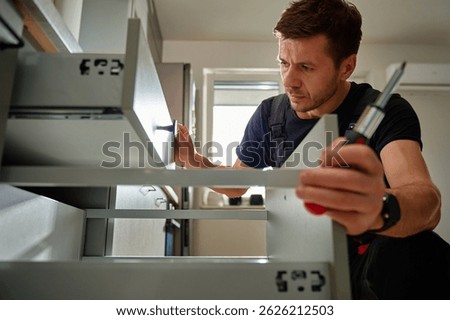 Man assembling kitchen drawer in modern cabinet using screwdriver. Handyman installing and adjusting drawer slide mechanism in contemporary kitchen. Furniture installation