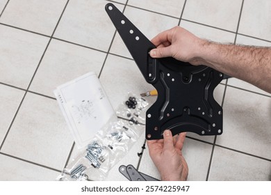 a man is assembling a TV bracket. He keeps instructions and is focused on the process. There are parts and tools nearby. Application: DIY, instructions, repair, TV accessories.
