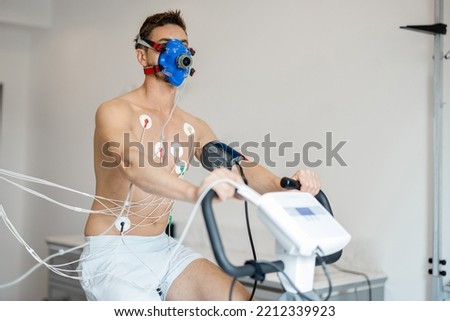 Man athlete with breath mask and electrodes training on bike simulator, examining his cardiovascular system at medical office