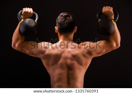 Man, athlete performs an exercise Kettlebell Military Press, posing standing with his back to the camera with naked torso in the studio. 
Young guy with weight, fitness model on the black background.