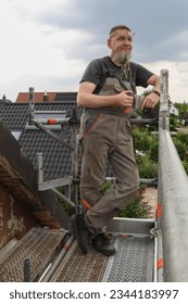 A man of athletic build with a beard in work clothes stands on the roof of a house on metal scaffolding and drinks coffee during a break from work
