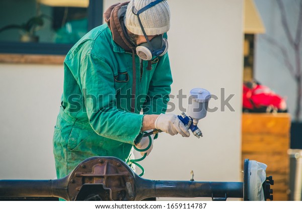 Man with automotive air\
spray gun in action during the restoration of a vintage car.\
Applying first base layer of paint onto an old big differential\
unit or rear axle.