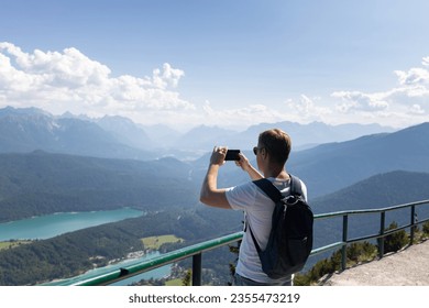 a man with a backpack behind his back photographs a beautiful landscape on his phone. A man standing on an observation deck overlooking mountains and lakes holds a phone in his hands.