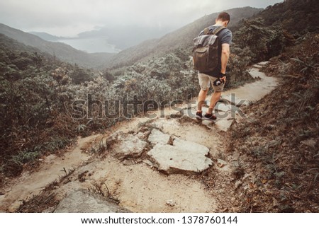 Man with backpack and camera is hiking by the stone path in the beautiful nature. There are fog and mountains at background.