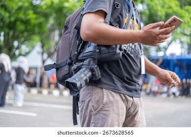 A man with a backpack and camera smokes while checking his smartphone during an outdoor street event in the city.