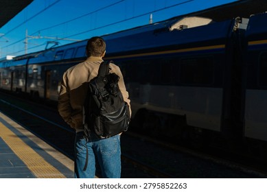 Man with backpack checking his smartwatch on a sunny train platform while waiting beside a passing train, showing travel, commute, schedule, and public transport.