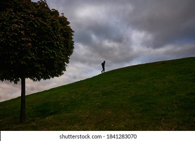 A Man With A Backpack Climbs Up A Hill Against A Sky With Dark Clouds