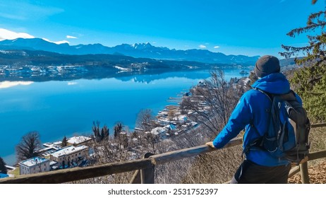 Man with backpack enjoying scenic view of alpine Lake Wörth surrounded by snow-capped mountains Karawanks, Carinthia, Austria, Europe. Alpine landscape reflected in calm water. Hohe Gloriette - Powered by Shutterstock