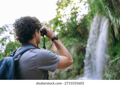 Man with backpack exploring nature, looking through binoculars at a waterfall. Concept of adventure, travel, and birdwatching