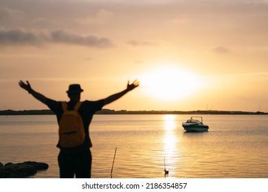 Man with backpack and hat opens his arms by the sea