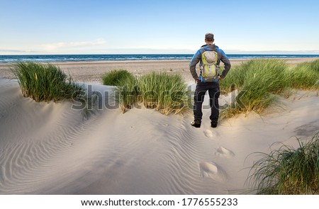 Man with Backpack hiking in beautiful windy coastal dune marram grass towards beach of North Sea in soft evening sunset sunlight. Skagen Nordstrand, Denmark. Skagerrak, Kattegat.