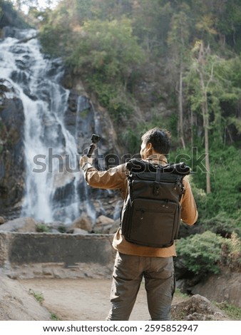 A man with backpack holding an action camera, infront of waterfall