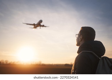 Man with backpack looking up to airplane landing at airport during beautiful sunset. 