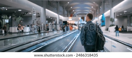 A man with a backpack moves to the gate on a travelator at the airport. A young tourist guy with hand luggage in a modern large airport terminal