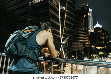 Man with backpack on a bridge railing observing the illuminated city skyline at night