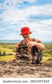 Man with backpack places tower of flat carne stones on rock outcrop with panoramic view of mountain valley. A man in moment of quiet ritual, grounded, absorbed in creating balance of stones