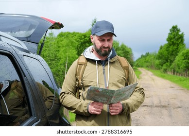 Man with backpack reading paper map beside car with open trunk on dirt road near green forest