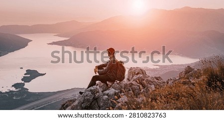 Man with backpack sitting on rocks, watching sunset over the calm waters of Bay of Kotor fjord landscape from a high mountain in Montenegro