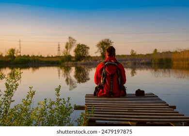 Man with a backpack sitting on a wooden pier, looking at a tranquil lake at sunset. Ideal for themes of travel, solitude, nature, reflection, and serenity. - Powered by Shutterstock