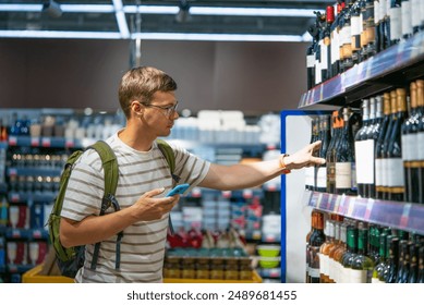 Man with backpack and smartphone selects bottle of wine from shelf in well-stocked liquor store. 
