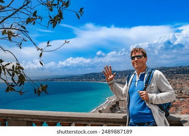 A man with a backpack is smiling and waving at a scenic coastal overlook against a blue sky and cityscape background. Nice, France. - Powered by Shutterstock