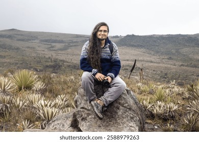 man with backpack smiling while hiking in a foggy mountainous landscape. Dressed in a warm sweater, surrounded by wild vegetation