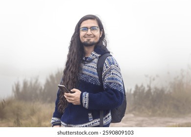 man with backpack smiling while hiking in a foggy mountainous landscape. Dressed in a warm sweater, surrounded by wild vegetation