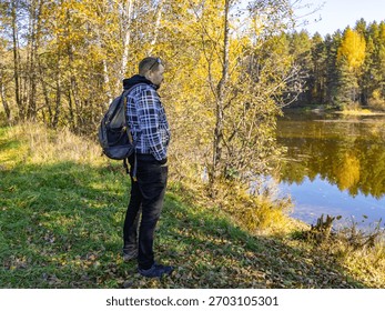 Man with a backpack is standing in a forest by a lake. The man is looking at the water, and the scene has a peaceful and serene mood