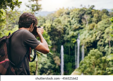 Man with backpack standing in front of waterfall and taking a photo. Male hiker photographing a beautiful water fall in forest - Powered by Shutterstock