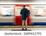 Man with backpack standing on a platform, watching a speeding subway train with motion blur in London. Mind the gap text visible