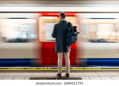 Man with backpack standing on a platform, watching a speeding subway train with motion blur in London. Mind the gap text visible