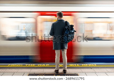 Man with backpack standing on a platform, watching a speeding subway train with motion blur in London. Mind the gap text visible