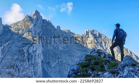 A man with a backpack standing in the shadow at the mountain peak and enjoying the view on higher, very steep peaks in front of him. Way to the Grimming in Austrian Alps. Freedom and exploration
