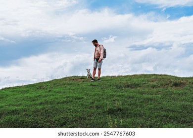 Man with backpack standing with small dog on green hilltop under cloudy blue sky - Powered by Shutterstock