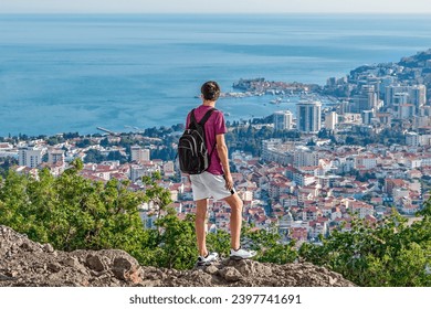 A man with a backpack and in summer clothes looks from the top of a mountain at the roofs of Budva and the Adriatic Sea in Montenegro. Solo hitchhiking in the Balkans - Powered by Shutterstock