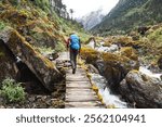 Man with backpack and trekking poles crossing mountain creek wooden bridge during Makalu Barun National Park trek in Nepal. Mountain hiking and active people concept image.