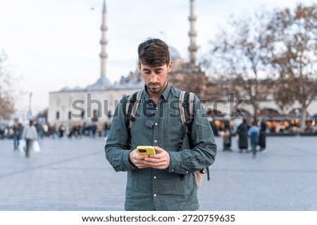 Man with backpack using smartphone in a busy public square with a mosque in the background in Istanbul