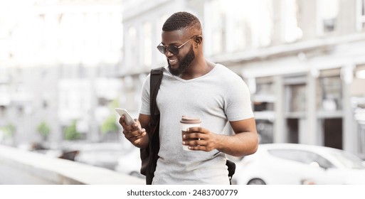 A man with a backpack walks down the street, looking at his phone and holding a coffee cup. - Powered by Shutterstock