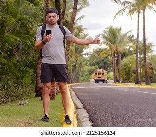 Man Backpacker Hitchhiker Stands By The Road With Thumb Up And Looking At Smartphone In His Hand. Single Traveler, Tourism, Hitchhiking Concept.