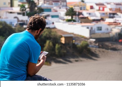 man from backside using cellular phone with internet connection and app. old town and beach in the background. summer time and leisure activity relaxing