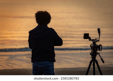 Man backview unrecognized lwith a camera on tripod next to him on the beach at sunrise