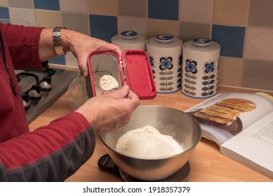 Man Baking, Putting Flour Into Weighing Scales. Making Pancakes With A Cook Book Open On The Work Top.