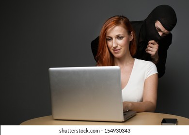 Man in a balaclava furtively watching an unsuspecting female office worker working on her laptop computer while corporate spying, stealing personal or business information or employee monitoring