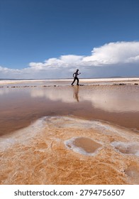 Man balancing on stone path across water pool at Salar de Uyuni salt flat in Bolivia, tourist exploring pink mineral water and white desert landscape under blue sky, South America travel